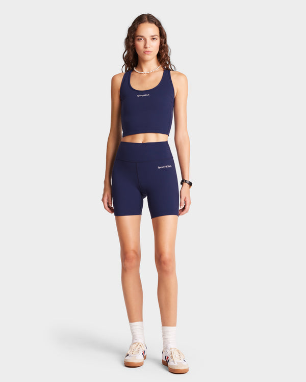 A woman in Serif Logo Biker Shorts, navy and white, stands against a plain white background, showcasing a luxurious athletic look.