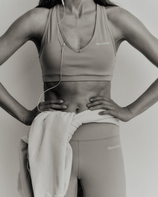 A person wearing a sports bra, Serif Logo Biker Shorts in Elephant/White, and a towel around the waist, enjoying music on earphones.