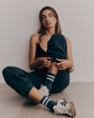 A person sits on the floor in a tank top, Serif Logo Sweatpant - Navy/White crafted from USA cotton, striped socks, and sneakers against a plain backdrop.