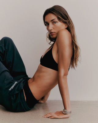 A person in black sportswear, featuring Navy/White Serif Logo Sweatpants made in the USA, sits against a wall with a relaxed posture, looking at the camera.