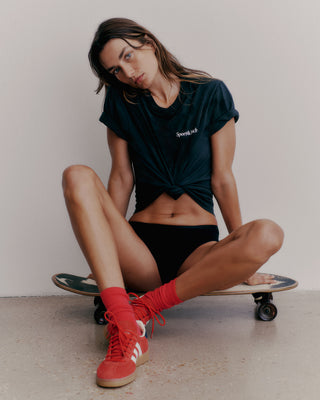 A woman confidently sits on a skateboard, wearing a Health is Wealth T-Shirt in navy/white and black shorts made from USA-grown cotton. Her red socks and sneakers provide a bold touch, all proudly crafted in the USA.