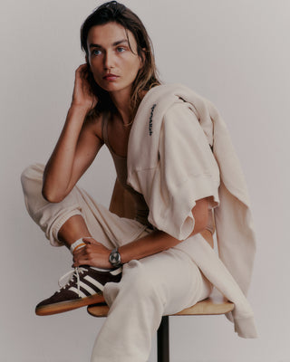 A person in a cream outfit, wearing the Serif Logo Sweatpant and accessorized with sneakers and a watch, sits on a stool. Their jacket—crafted in the USA—draped over their shoulder, exudes effortless style in this unisex ensemble.