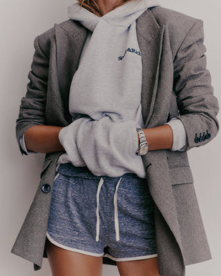 A person in a Serif Logo Hoodie - Heather Gray/Navy, paired with blue shorts and hands in pockets, showcases a unisex style. Made from USA grown cotton, it highlights quality craftsmanship made in the USA.