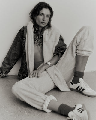 A person sits against a wall, wearing a denim jacket made from USA-grown cotton, paired with Serif Logo Sweatpant in Heather Gray/Navy and sneakers.
