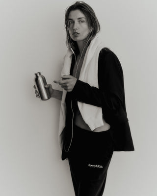 A person with a towel over their shoulders, holding a metal bottle and wearing headphones, is dressed in a jacket and pre-shrunk Serif Logo Sweatpant in Faded Black/White.