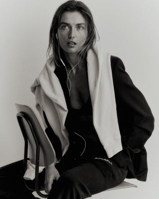 A woman sits wearing a pre-shrunk jacket and headphones, draped with a hoodie, in a monochrome setting. She pairs the look with Serif Logo Sweatpants in faded black/white casual elegance.