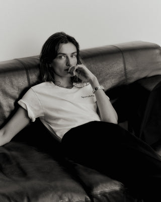 A person in the Health is Wealth T-Shirt (Cream/Navy), made in the USA with 100% cotton, lounges on a leather couch, gazing thoughtfully at the camera in their dark pants.