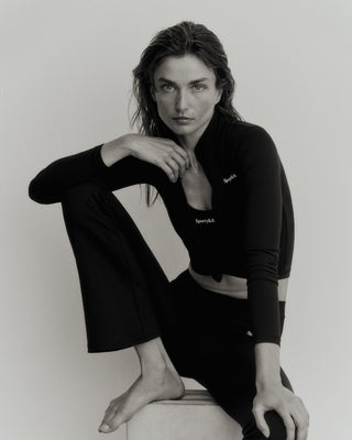 Person in a Serif Logo Sports Jacket - Black/White, fabric of the highest quality, long-sleeve with zipped closure, sitting and looking at the camera with one knee raised in a black and white image.