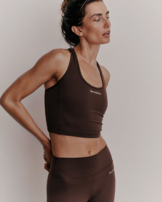 In a sleek Serif Logo Sports Tank in Chocolate/White, the woman poses gracefully against a light background, her gaze thoughtfully directed to the side.