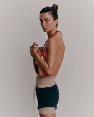 A person with wet hair holds sandals, wearing a red halter top and Serif Logo Biker Short in Elephant/White, gazing forward against a plain backdrop.