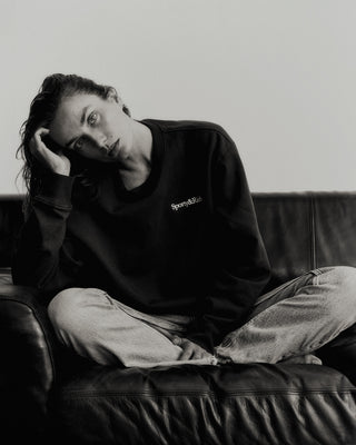 A person sits cross-legged on a leather couch, wearing the Serif Logo Crewneck in Faded Black/White, crafted from USA-grown cotton, along with jeans, resting their head on their hand.