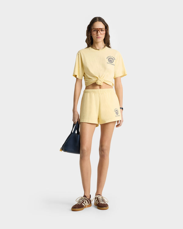 Woman in yellow knotted 100% cotton T-shirt and California Emblem Disco Short in Almond/Navy with elastic waistband, holding a black handbag, wearing glasses and brown sneakers.