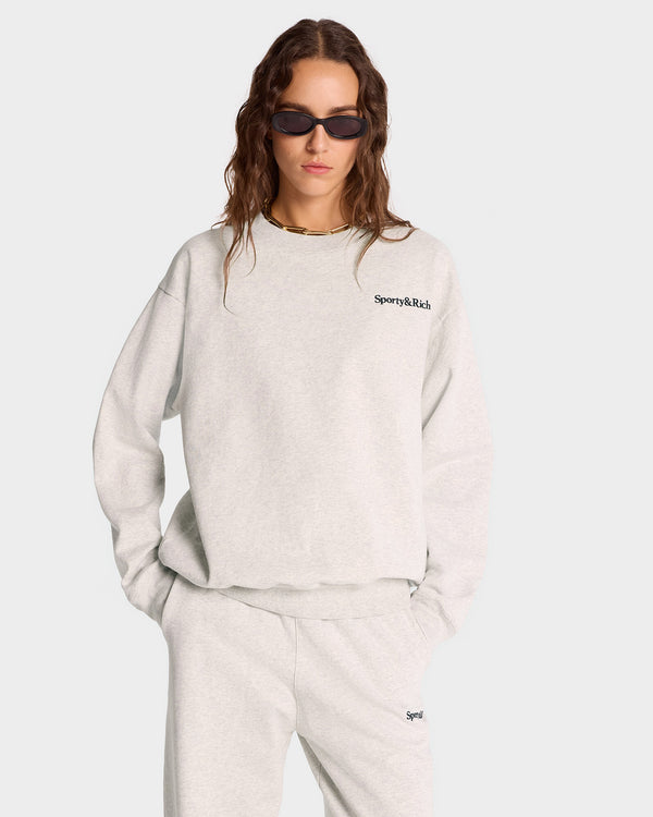 A person is wearing sunglasses with a stylish gray Serif Logo Crewneck - Heather Gray/Navy and matching pants, crafted from USA-grown cotton, standing against a plain background.