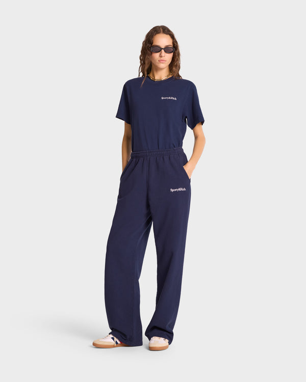 Person wearing the Serif Logo Straight Leg Sweatpant in Navy/White, made from USA-grown cotton, a navy blue t-shirt, sunglasses, and white sneakers against a plain white background.