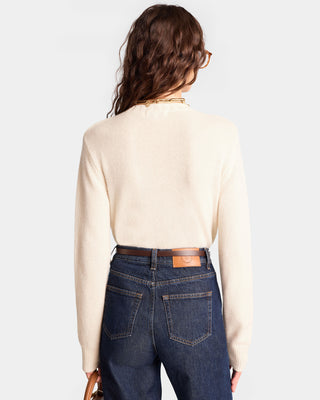 Model is wearing The Carlyle Crest Cashmere Crewneck in Cream/Forest, featuring ribbed edges and a cropped fit, paired with jeans. They face away while holding a small brown purse.