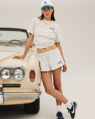 In a studio, a woman in the Dodgers Serif Disco Short in Heather Gray/Navy leans against a vintage beige car, wearing sneakers and a cap.