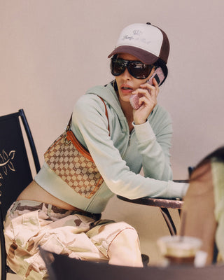 Person sitting at a table wearing a light green hoodie, camouflage pants, sunglasses, and a cap, with a patterned bag.