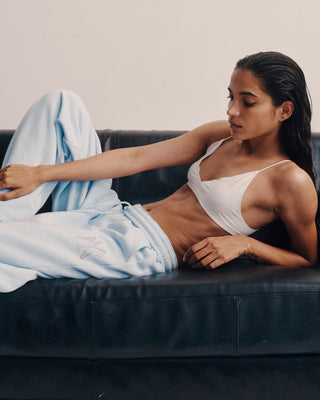 Woman in a white tank top and light blue sweatpants sitting on a black leather couch.