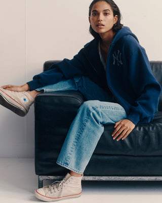 Person wearing a blue jacket with a logo, light blue jeans, and white sneakers sitting on a black leather couch.