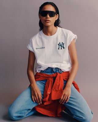 Person wearing a white t-shirt with logos, blue jeans, and a red scarf sitting on the ground.