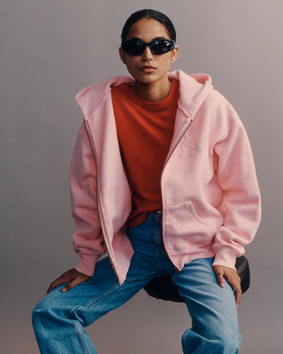 Person wearing a pink hoodie, orange shirt, and blue jeans sitting on a chair against a gray background.