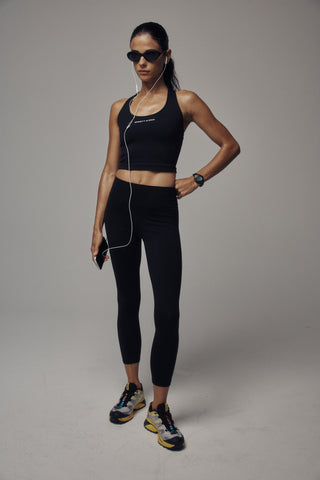 Woman in a black Fran Bold Logo Sports Tank with a supportive fit, sunglasses, and headphones stands holding a phone, posing confidently.