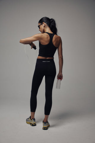 Woman in a black Fran Bold Logo Sports Tank with a supportive fit, sunglasses, and headphones stands holding a phone, posing confidently.