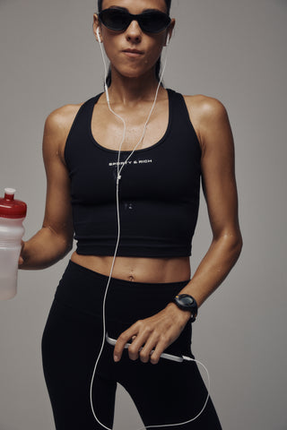 Woman in a black Fran Bold Logo Sports Tank with a supportive fit, sunglasses, and headphones stands holding a phone, posing confidently.
