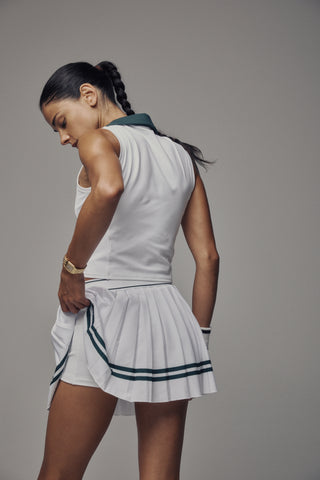 The Ally Serif Logo Pleated Skirt - White/Forest features crisp pleats and green forest stripes near the hem, with Sporty & Rich text on the elastic waistband and comfortable built-in shorts for ease of movement.