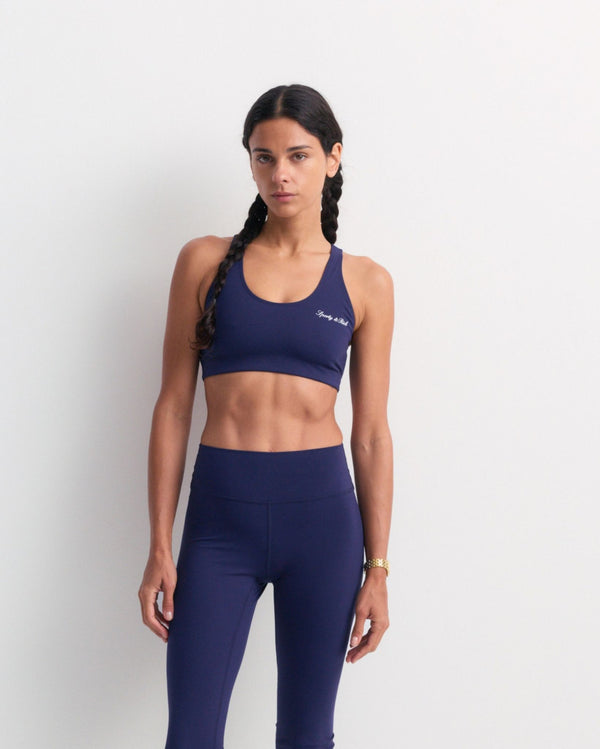 A woman stands against a plain white wall in the Billie Signature Logo Sports Bra - Dark Navy, arms at her sides and ready for high-impact workouts, supported by its racerback design.