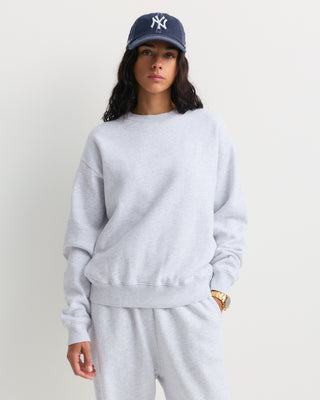 The Eeva Serif Logo Embroidered Crewneck in Heather Gray features a classic crewneck design with long sleeves and subtle white embroidered text on the left chest, making it perfect for fans of vintage-style sweatshirts.