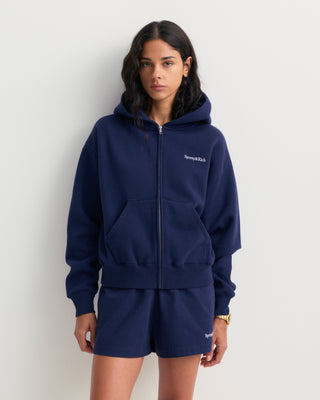 Emaro Serif Logo Embroidered Zip Hoodie in Dark Navy, laid flat. This relaxed fit hoodie features front pockets, an embroidered Sporty & Rich logo on the left chest, and an ultra-soft feel for everyday comfort.