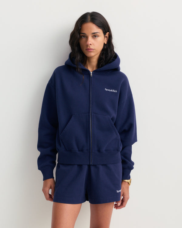 Emaro Serif Logo Embroidered Zip Hoodie in Dark Navy, laid flat. This relaxed fit hoodie features front pockets, an embroidered Sporty & Rich logo on the left chest, and an ultra-soft feel for everyday comfort.