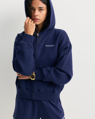Emaro Serif Logo Embroidered Zip Hoodie in Dark Navy, laid flat. This relaxed fit hoodie features front pockets, an embroidered Sporty & Rich logo on the left chest, and an ultra-soft feel for everyday comfort.