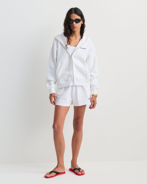 The Lucy Serif Logo Embroidered Disco Short – White is crafted from 100% cotton, featuring an elastic waistband and the Sporty & Rich logo embroidered on the lower left leg, making it perfect as a sporty lounge short. Shown here against a gray background.