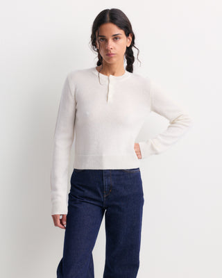 A woman modeling a cream-colored long-sleeve knit sweater with a short button placket, paired with dark blue jeans, against a plain white studio background.