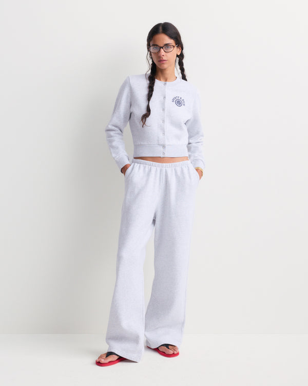 Woman in glasses wearing a relaxed, oversized heather gray Besma Serif Logo Embroidered Baggy Sweatpant and matching top with red flip-flops, standing against a white wall.