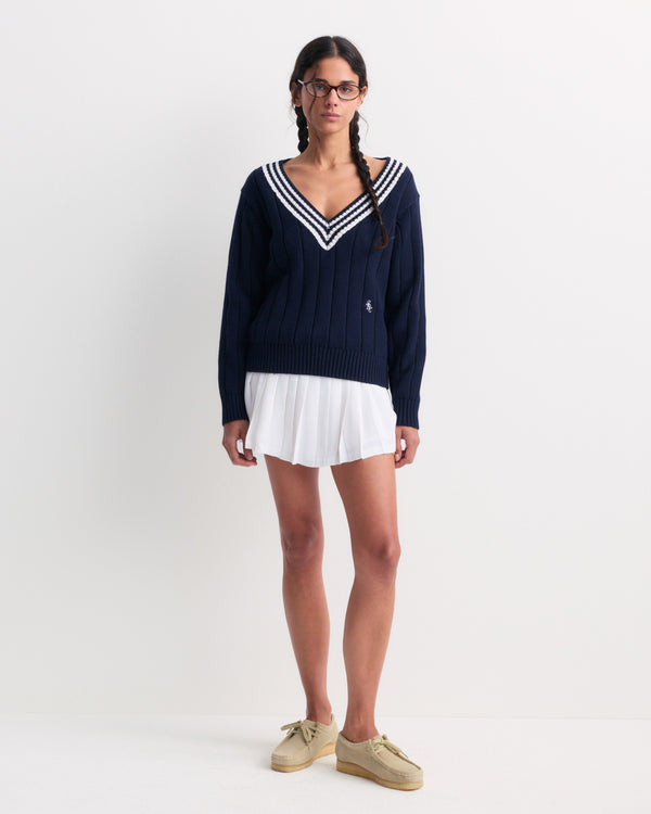 A woman wearing glasses, beige shoes, a navy V-neck sweater, and the Abigail Signature Logo Abigail Skirt in Off White stands against a white wall.