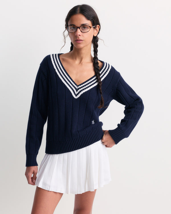 A woman modeling a navy blue ribbed knit sweater with a white and navy striped V-neck collar, paired with a white pleated skirt, against a plain light gray studio background.