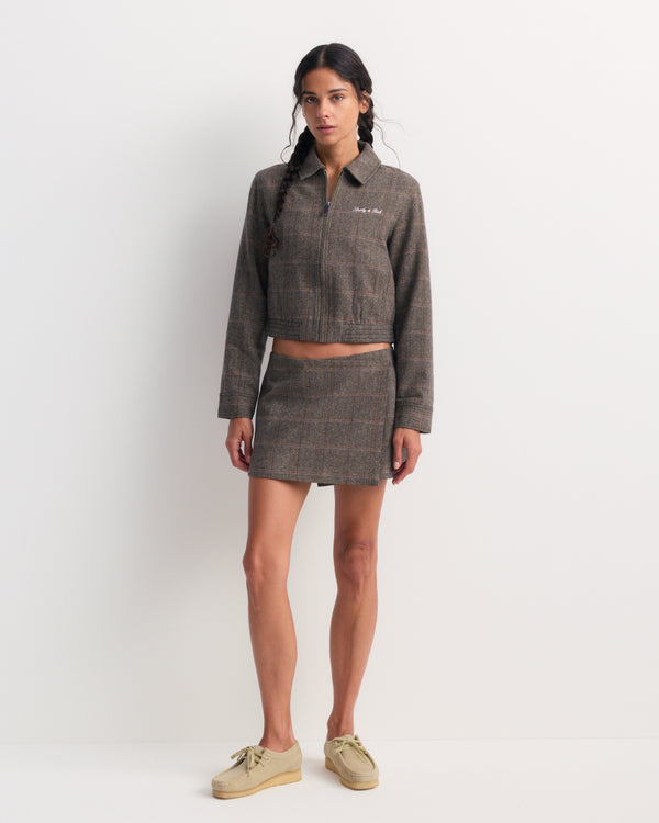 Woman wearing the Astrid Signature Logo Folding Skirt in brown check with a cropped plaid zip-up jacket, standing against a plain white wall.