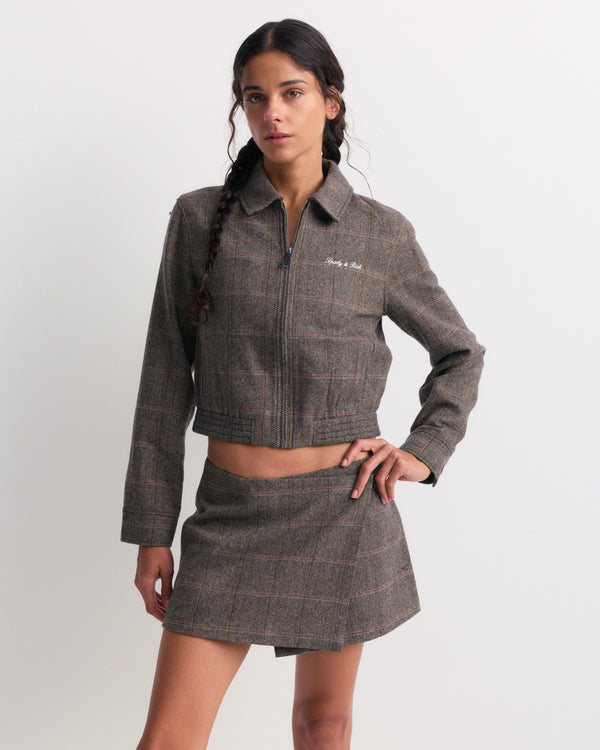 A woman models the Gyl Signature Logo Smart Zip Jacket in brown check with a matching skirt, standing with one hand on her hip against a plain background.