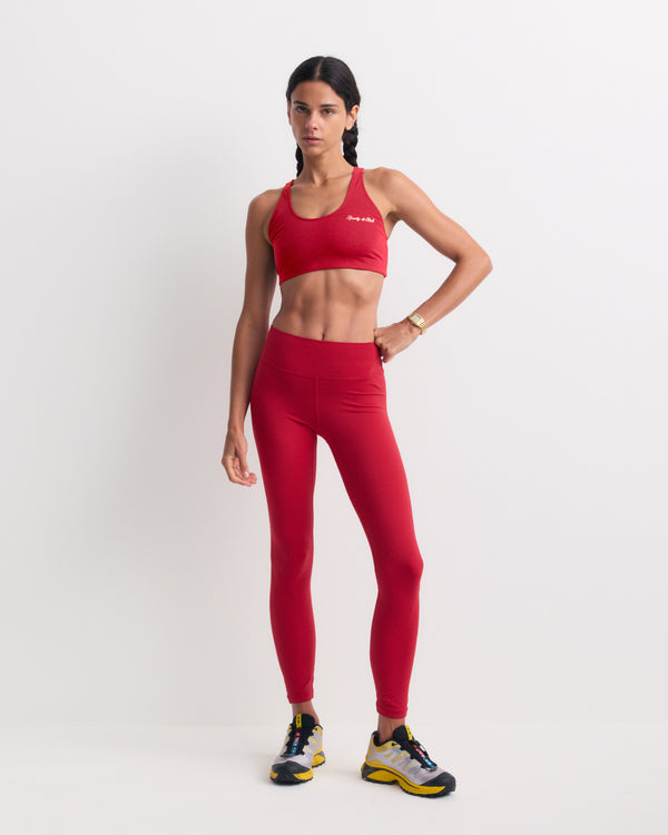 A woman stands against a white background wearing the Bonnie Signature Logo Legging in Ruby, paired with a matching sports bra and yellow and black sneakers. These opaque workout leggings deliver a supportive fit for any fitness routine.