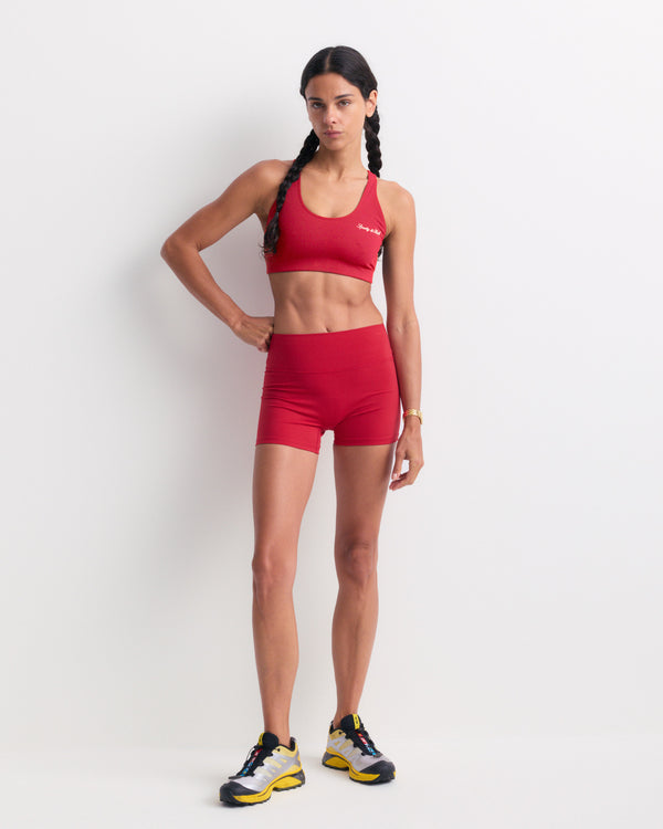 A woman stands against a plain white background, her hair in braids and wearing sneakers, dressed in a red sports bra and the Blaire Signature Logo Mini Biker Short in Ruby. The high-waist fit and super soft fabric of these shorts ensure comfort while she moves.