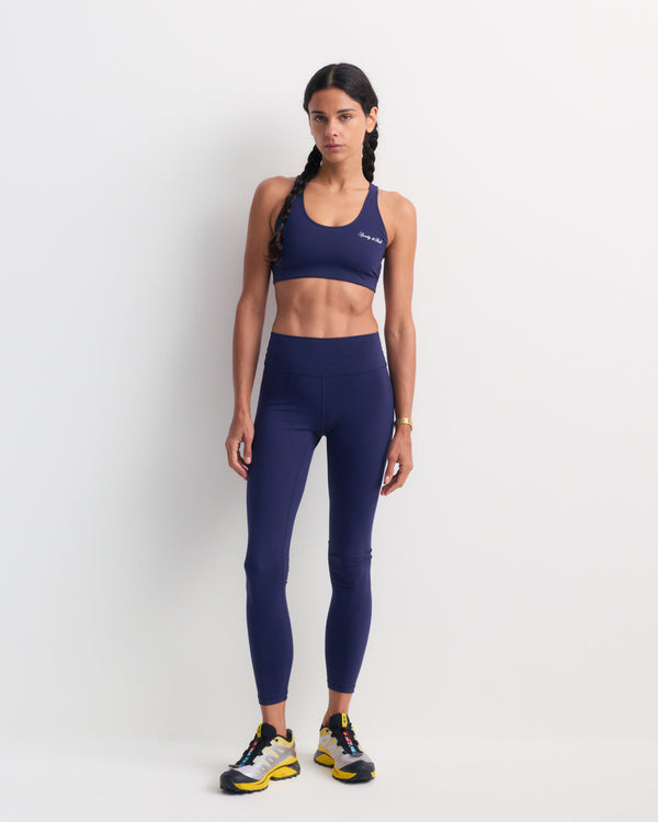 A woman is wearing the Bonnie Signature Logo Legging in Dark Navy with a supportive fit sports bra and sneakers, standing against a plain white background.