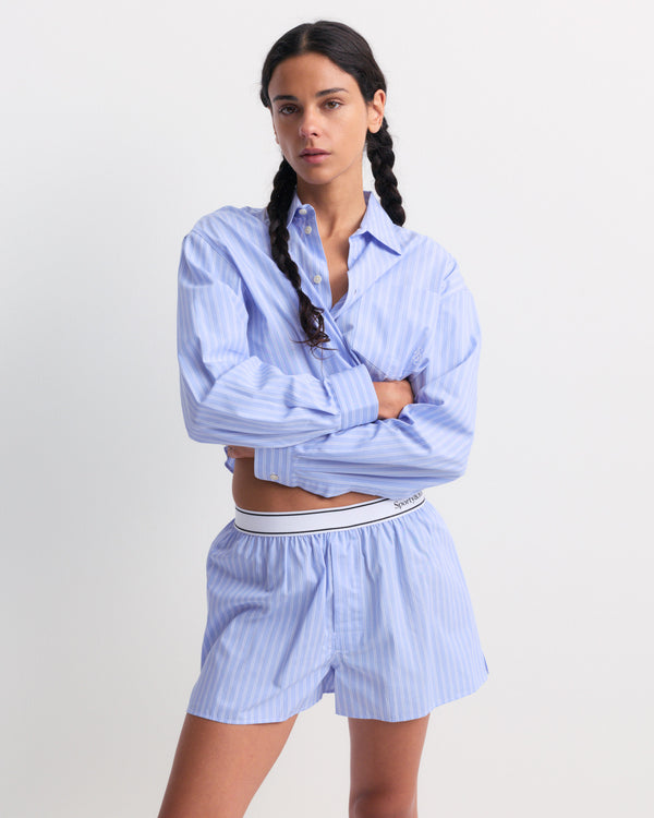 The Coco SRC Oversized Shirt - Blue Striped features a light blue, long-sleeve, button-up design with white vertical stripes and a chest pocket. Crafted from breathable poplin for a relaxed fit, it’s shown laid flat.