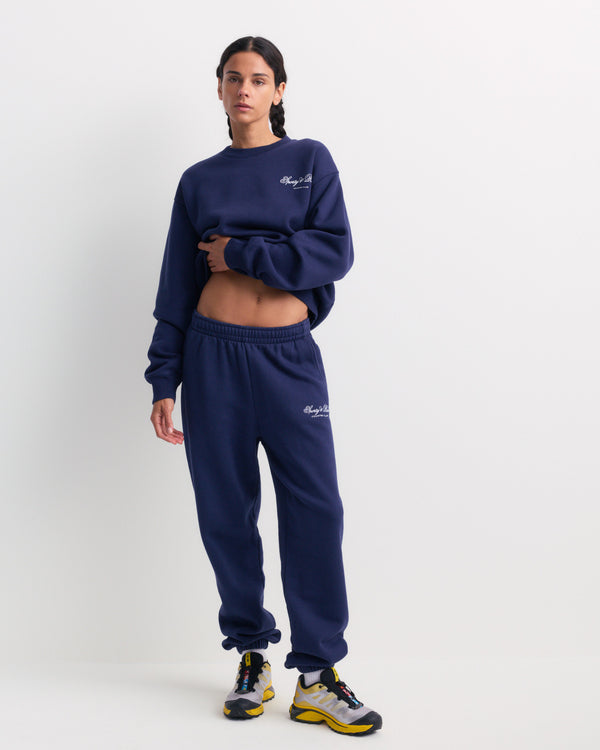 A woman in the Brianno Club Script Jogger in Dark Navy, styled with a matching sweatshirt and yellow sneakers, stands against a plain white background.