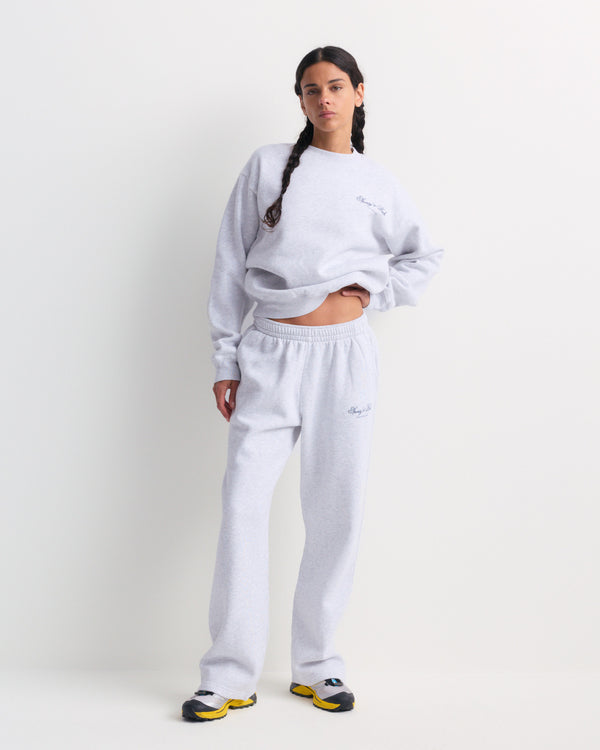 A woman wearing the Blakely Club Script Wide Sweatpant in Heather Gray stands against a plain white background with her hands in her pockets, paired with yellow sneakers.
