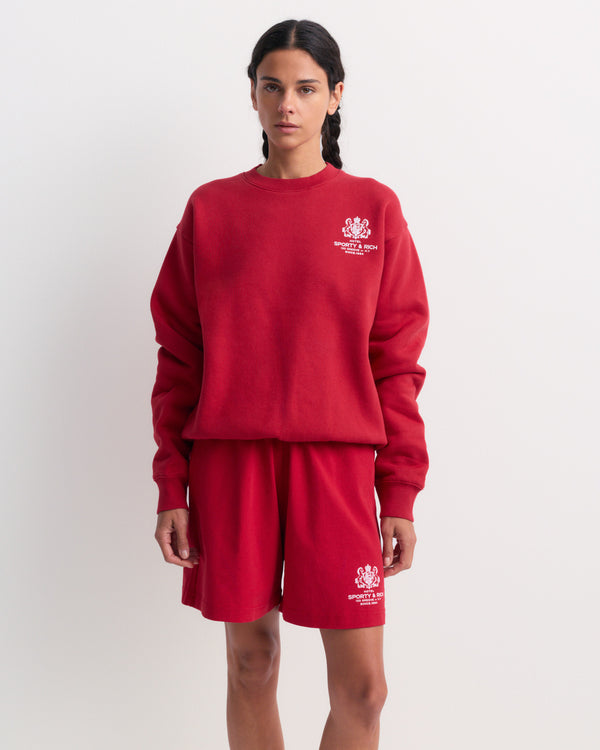 The Emmett Sr Hotel Crewneck - Ruby is a red crewneck sweatshirt inspired by vintage styles, featuring “Sporty & Rich” white logo and text on the left chest area.