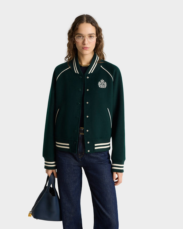 Woman wearing glasses, the H&W Crest Double Face Varsity Jacket in Forest/White with leather details, dark jeans, and holding a blue handbag, standing against a plain background.