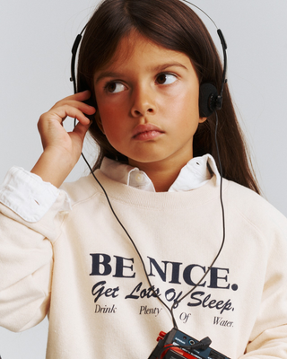 A child wearing a cream Kenza Be Nice Kids Crewneck printed with: BE NICE. Get Lots of Sleep. Drink Plenty Of Water.
